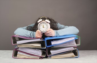 Exhausted tired woman with alarm clock sleeping on a pile of file folders, burnout, stress and