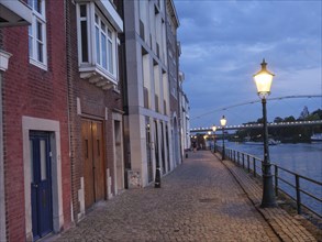Riverside street at dusk with illuminated lanterns and houses along the pavement, Maastricht,