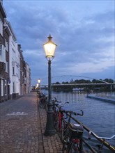 Street scene on the riverbank at dusk with bicycles and lanterns, bridge in the background,