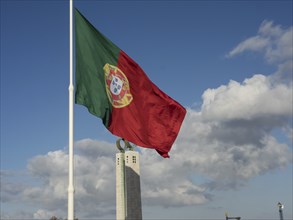 A Portuguese flag flies in front of a tall monument under a sky with scattered clouds, Lisbon,