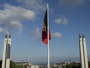 A large Portuguese flag flies against a blue sky with some clouds and buildings in the background,