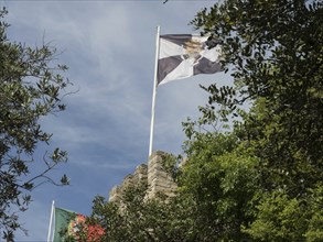 Two flags waving on a castle wall, surrounded by trees under a blue sky, Lisbon, portugal