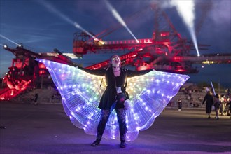 Festival visitor with a bright dress dances in front of an excavator at the Melt Festival in