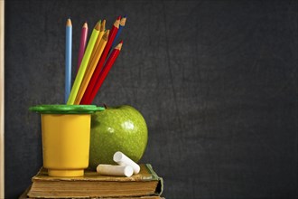 Multicolor pencil, chalk and green apple on old textbook against blackboard in class. School