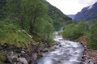 A calm river flows through a green mountain landscape, surrounded by trees and cloudy skies, Olden,