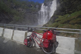 A packed bicycle stands on a road in front of an impressive waterfall in a mountainous landscape,