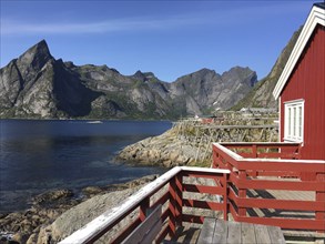 Wooden railing by the water with a red hut in the foreground, majestic mountains in the background,