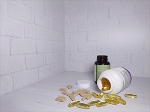 Two bottles of vitamins with spilled pills on a white surface against a white brick wall