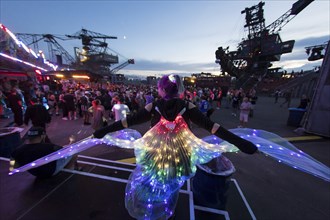 Festival visitor with a bright dress between excavators in the evening at the Melt Festival in