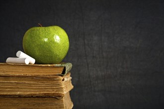 Chalk and green apple on old textbook against blackboard in class. School concept