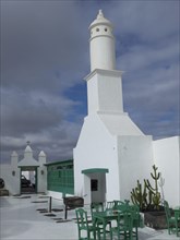 White lighthouse with buildings, green chairs and tables under a cloudy sky in Lanzarote, arrecife,