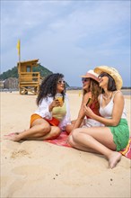 Three women sitting on a beach, one of them holding a drink. Scene is relaxed and carefree