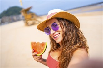 Selfie of a caucasian woman enjoying summer on the beach on vacation with hat and a watermelon