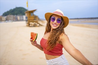 A woman is smiling and holding a watermelon slice. She is wearing a straw hat and sunglasses