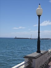 Coastal view of the calm sea with a lantern in the foreground and a harbour in the background under