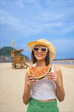 A woman is holding a watermelon on a beach. She is smiling and wearing a straw hat