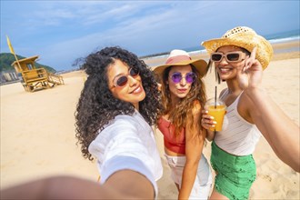 Three women are posing for a picture on the beach, with one of them holding a drink. Scene is