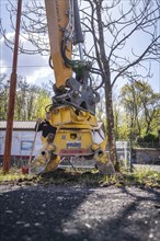 An excavator with grab arm stands in front of a bare tree and a building, demolition site,