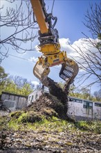 An excavator with a grab arm lifts earth while the sun is shining and trees and buildings can be