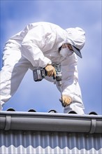 Worker in a white protective suit using a drilling machine on a roof under a blue sky, demolition