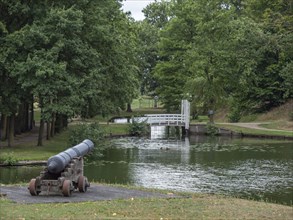 A historic cannon stands in a park on the banks of a river, surrounded by green trees and a small