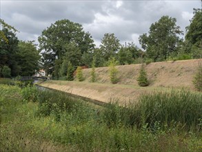 Natural landscape with a river bank surrounded by dense vegetation and trees under a cloudy sky,