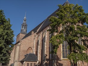 Brick church with tower and high windows, surrounded by trees under a blue sky, groenlo,