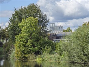 Shore of a lake with dense trees and modern buildings in the background, with clouds in the sky in