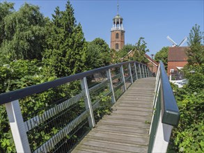 Wooden bridge leads to a church tower and a windmill, surrounded by trees under a blue sky, Ditzum,