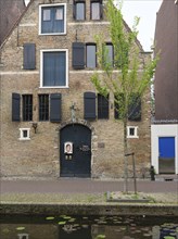 A historic brick building with black shutters, green trees and a quiet canal in the foreground,