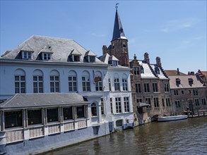 Historic buildings with sloping roofs and a tower along a quiet canal under a clear sky, Bruges,