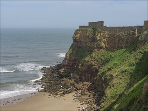 Coastal area with steep cliff jutting into the sea and historic remains above and a stretch of