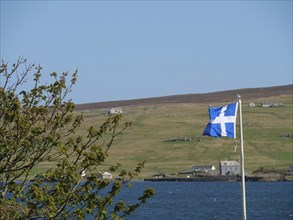 A Scottish flag flies on the coast with green hills and scattered houses in the background,
