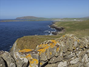 Stone wall with seaweed in the foreground, behind it sea and green coastal landscape under a blue