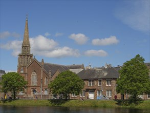 Church tower and urban buildings along a river with trees and blue sky with clouds, inverness,