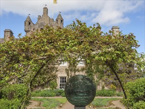 Castle with towers in the background, an archway of plants and a sculpture in a well-tended garden,