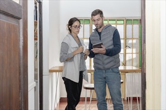 Two co-workers are seen discussing while having coffee. The man is holding a tablet and showing