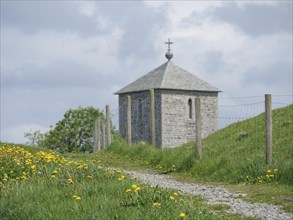 Stone church with picket fence on a green path, surrounded by yellow flowers and cloudy sky,