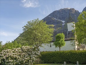 White church with steep roof surrounded by lush greenery and flowering shrubs, surrounded by