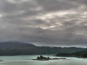 Small island in calm water with melancholic cloudy sky and mountains in the background, Eidfjörd,