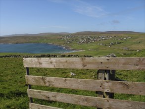 Wooden fence overlooking a coastline and a village surrounded by green hills under a blue sky with