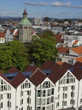 View of a city with a tower, modern buildings and a green treetop under a cloudy sky, Stavanger,