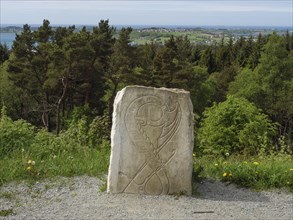 Stone with engraving in front of a green forest and sea view under a slightly cloudy sky,