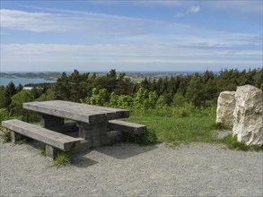 Wooden table and bench on a gravel path with a view of the forest and sea under a slightly cloudy