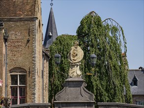 Statue in the foreground in front of historic brick buildings, a church with spire and gardens,