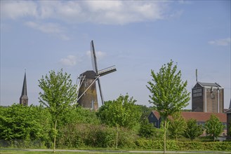 Windmill and church tower rise above the trees and buildings of a village under a clear sky,