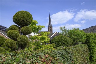 Church tower rises above neatly trimmed bushes and garden plants of a village under a clear sky,