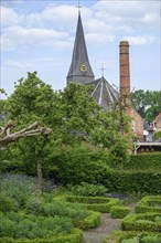 A church with a high tower next to an old chimney, surrounded by a well-tended garden and green