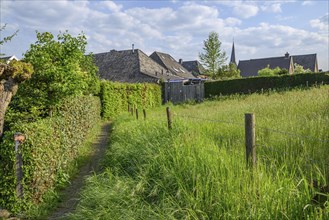 A narrow path leads through a green meadow along a row of houses, surrounded by green hedges, in a