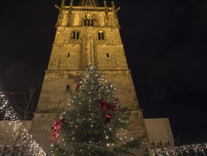 A tall church tower with a festively decorated Christmas tree full of fairy lights below, all at